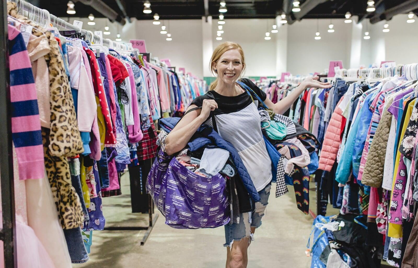 Mom wears baby and holds toddler boy clothes in each hand. Second mom shops behind her from the racks of boys clothes.